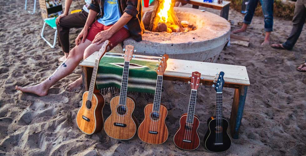 Na areia da praia, encostados num banco, cinco ukuleles de tamanhos diversos. Atrás uma fogueira, pessoas e cadeiras de praia. 