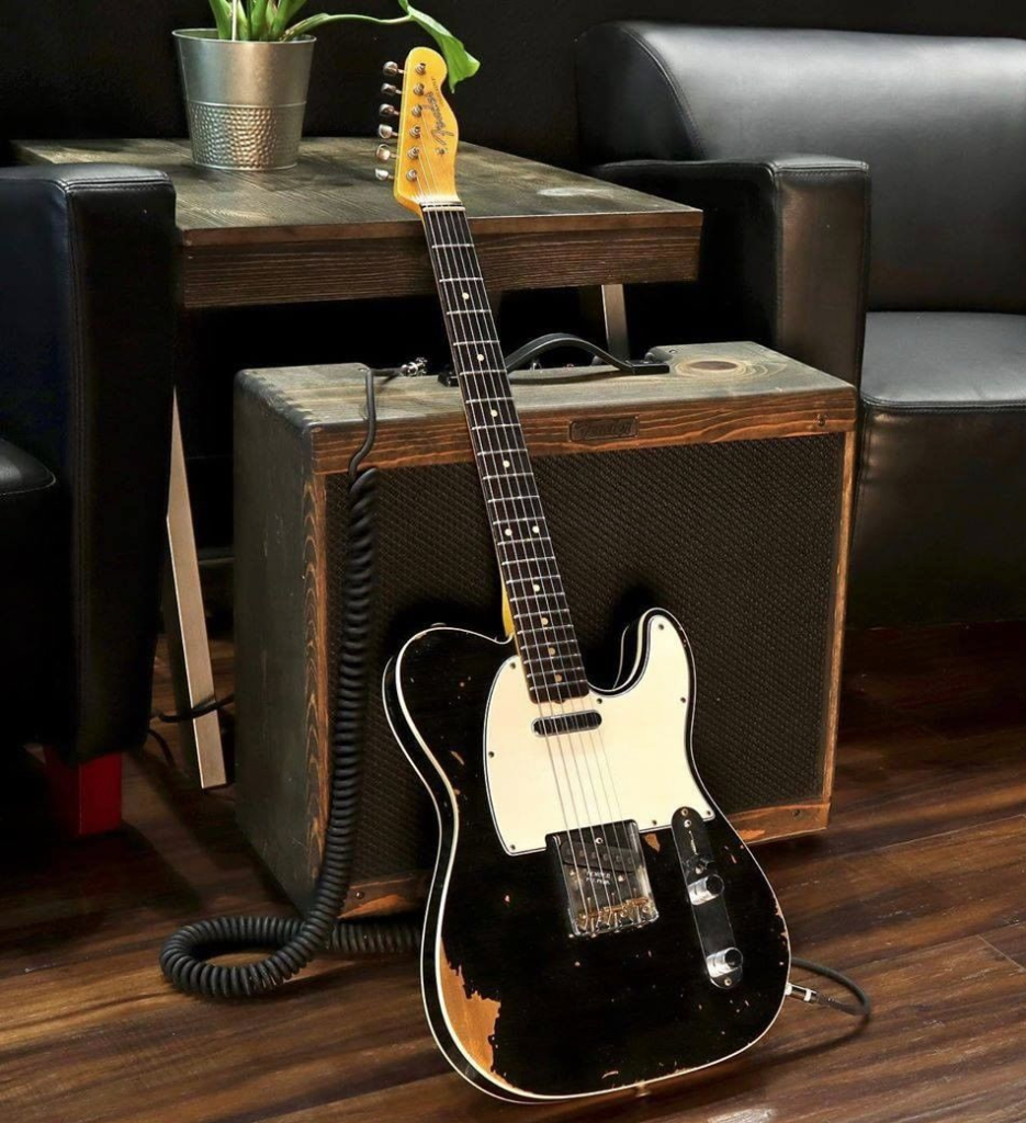 Guitarra Telecaster preta no chão de madeira encostada num amplificador Vintage. Em volta sofás pretos de couro. 